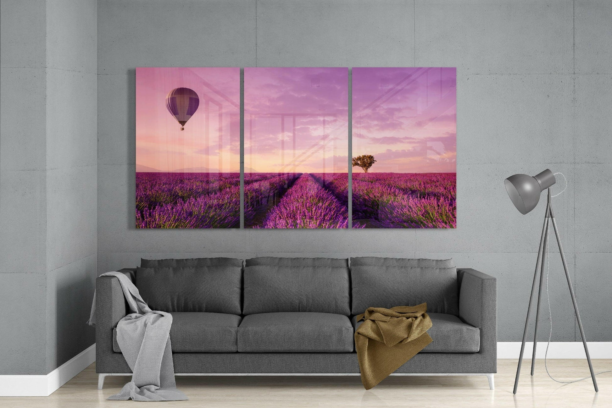 Three-panel lavender field glass art hung above gray sofa, depicting purple flowers, hot air balloon, and pink sunset sky.