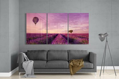 Three-panel lavender field glass art hung above gray sofa, depicting purple flowers, hot air balloon, and pink sunset sky.