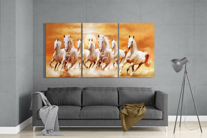 Three-panel glass art of white horses galloping through golden clouds displayed above a gray sofa in a modern living room.