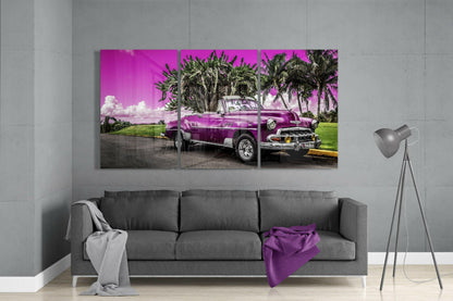 Three-panel glass art depicting a purple vintage car parked under palm trees against a vibrant magenta sky, displayed above a gray sofa.
