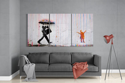 Three-panel glass wall art depicting a figure with colorful umbrella and child in rain, displayed above gray sofa in modern interior.