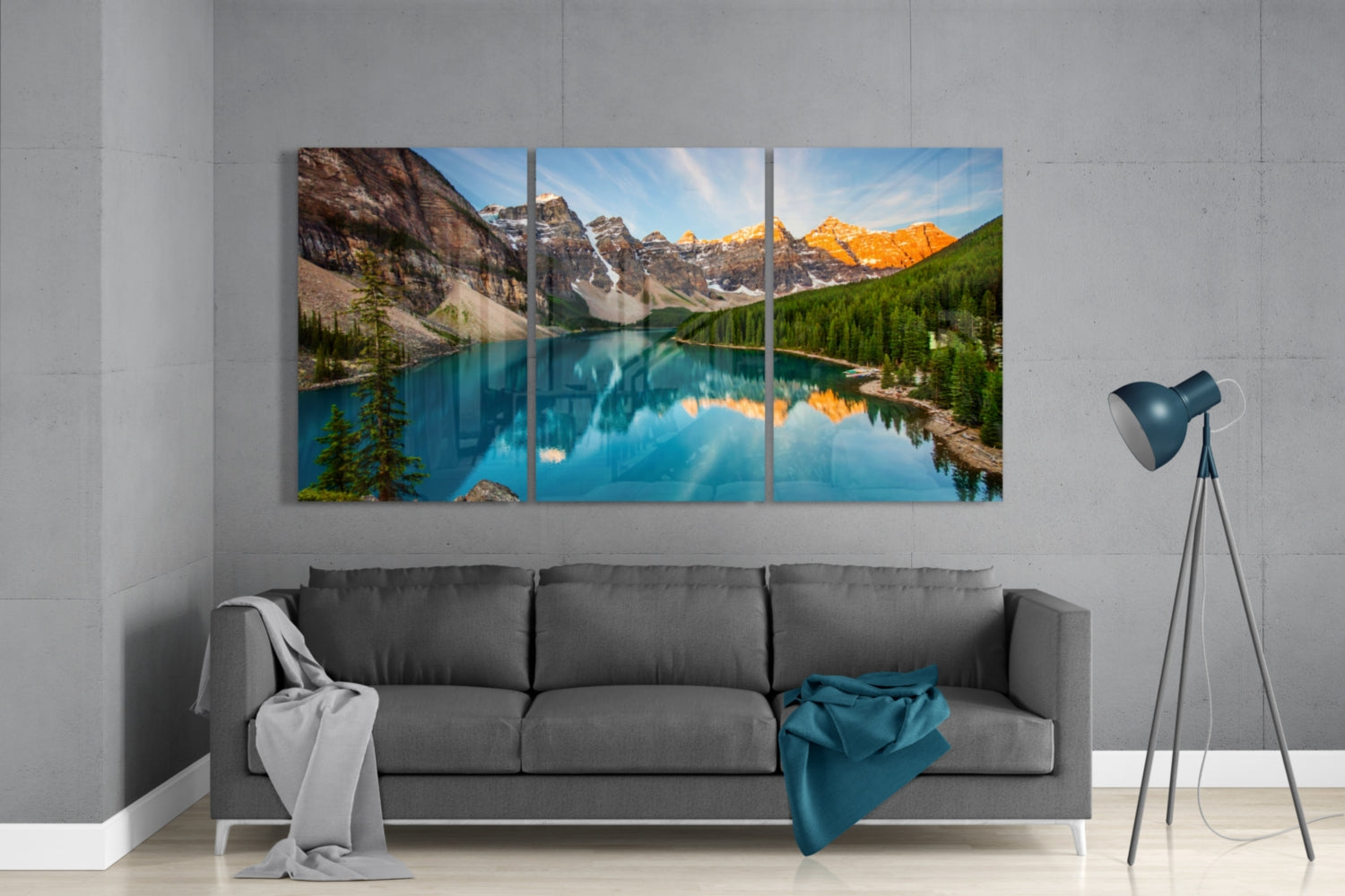 Three-panel glass wall art depicting a turquoise alpine lake surrounded by snow-capped mountains and evergreen forest, displayed above a gray sofa in a modern living room.
