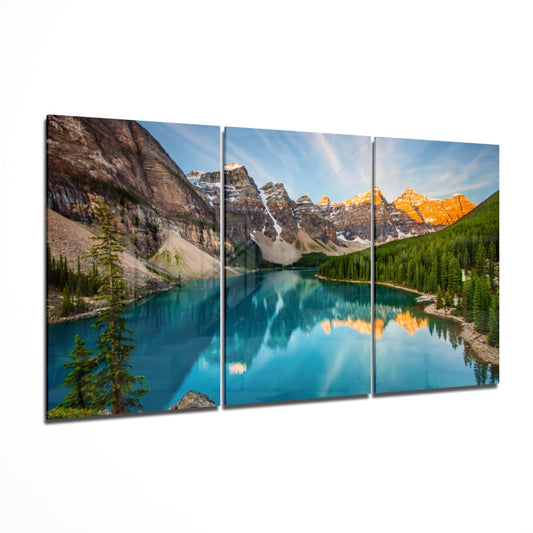 Three-panel glass wall art displaying a turquoise alpine lake surrounded by snow-capped mountains and evergreen forests under blue sky.