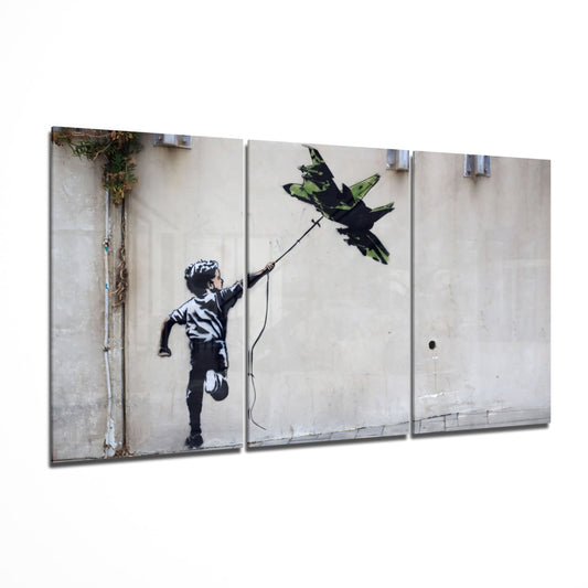 Three-panel glass wall art displaying stenciled black silhouette of child reaching toward green military jet on concrete wall.