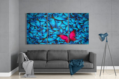 Three-panel glass wall art displaying blue butterflies with one red butterfly, mounted above gray sofa in modern living room.