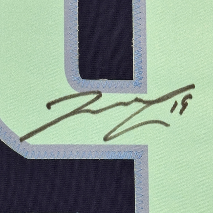Close-up of handwritten signature in dark ink on light green fabric with blue stitched border framing.