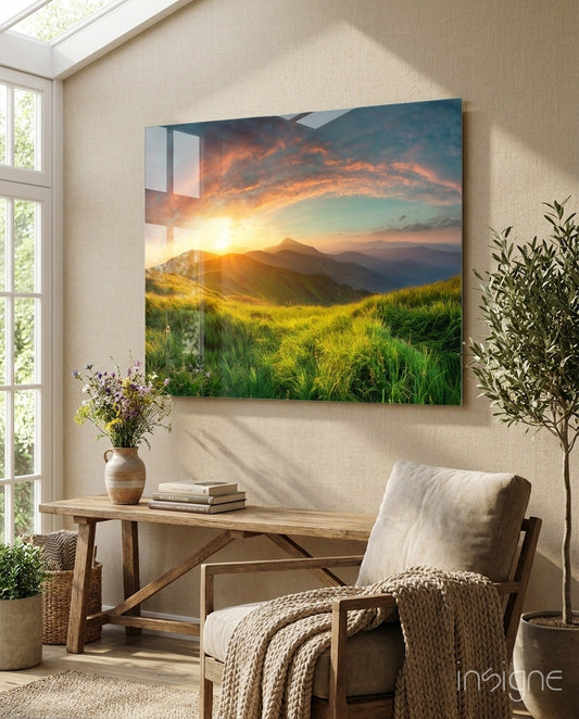 Glass wall art depicting a sunset over green hillside landscape displayed above wooden console table in bright room with plants and chair.