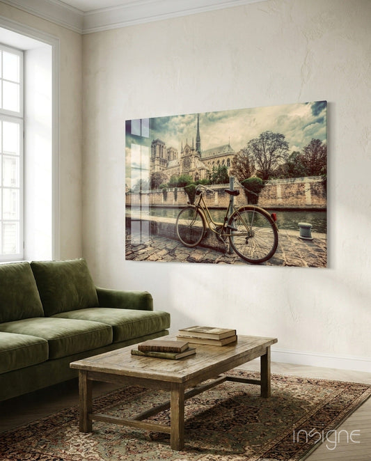 Glass wall art depicting a sepia-toned bicycle by a riverside cathedral, displayed above a green sofa in a living room.