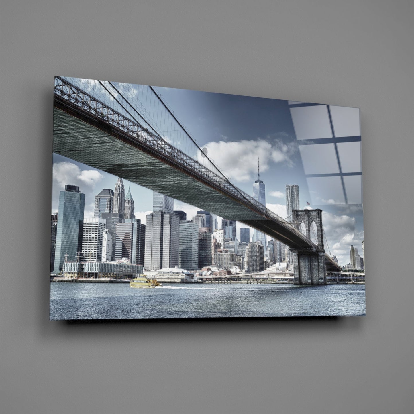 Glass wall art mounted on gray wall displaying Brooklyn Bridge spanning East River with Manhattan skyline and blue sky with white clouds.