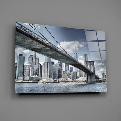 Glass wall art mounted on gray wall displaying Brooklyn Bridge spanning East River with Manhattan skyline and blue sky with white clouds.