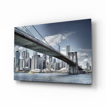 Glass wall art displaying Manhattan Bridge spanning over blue East River with NYC skyline and skyscrapers beneath cloudy sky.