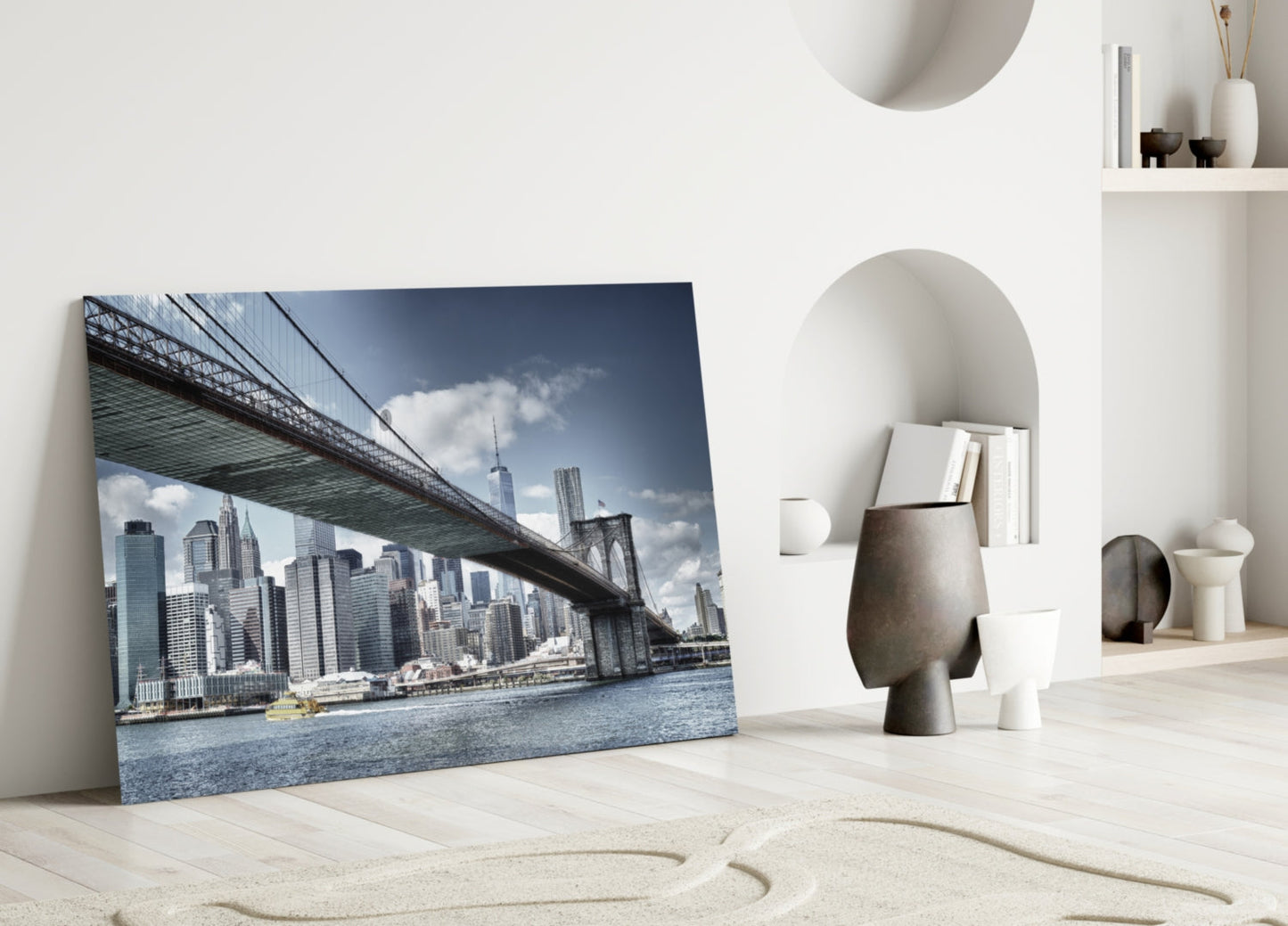 Glass wall art leaning against white wall displays Brooklyn Bridge spanning Manhattan skyline with blue water and cloudy sky.