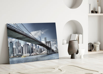 Glass wall art leaning against white wall displays Brooklyn Bridge spanning Manhattan skyline with blue water and cloudy sky.