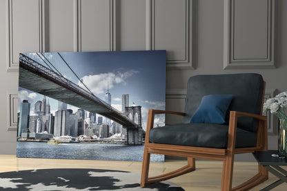 Manhattan Bridge glass wall art displaying Brooklyn Bridge spanning East River with Manhattan skyline, positioned in modern living room beside wooden chair.
