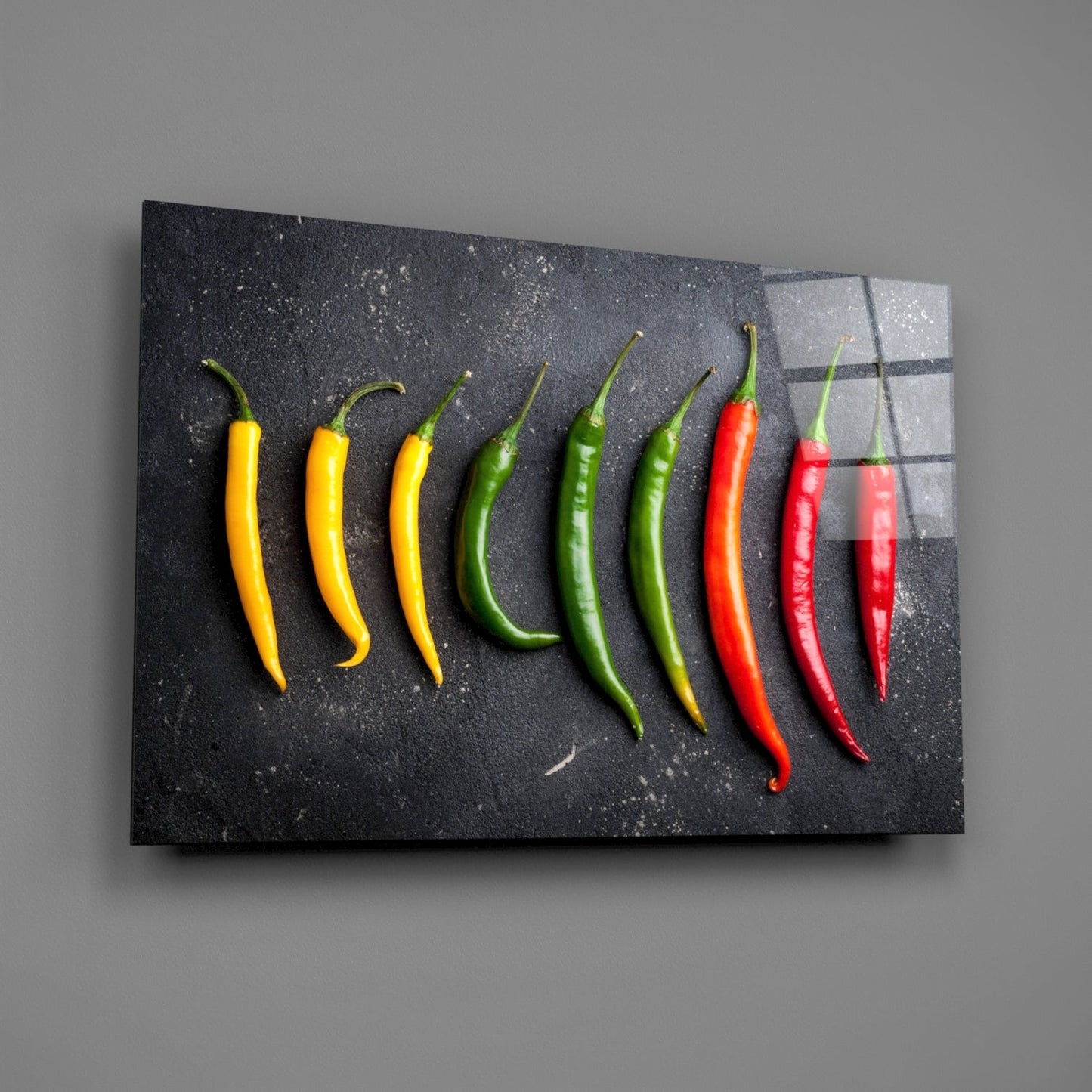 Nine chili peppers arranged in gradient from yellow to red on a dark textured glass panel mounted on gray wall.