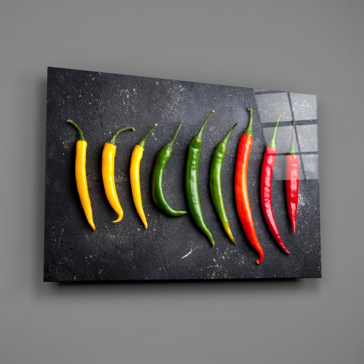 Nine chili peppers arranged in gradient from yellow to red on a dark textured glass panel mounted on gray wall.