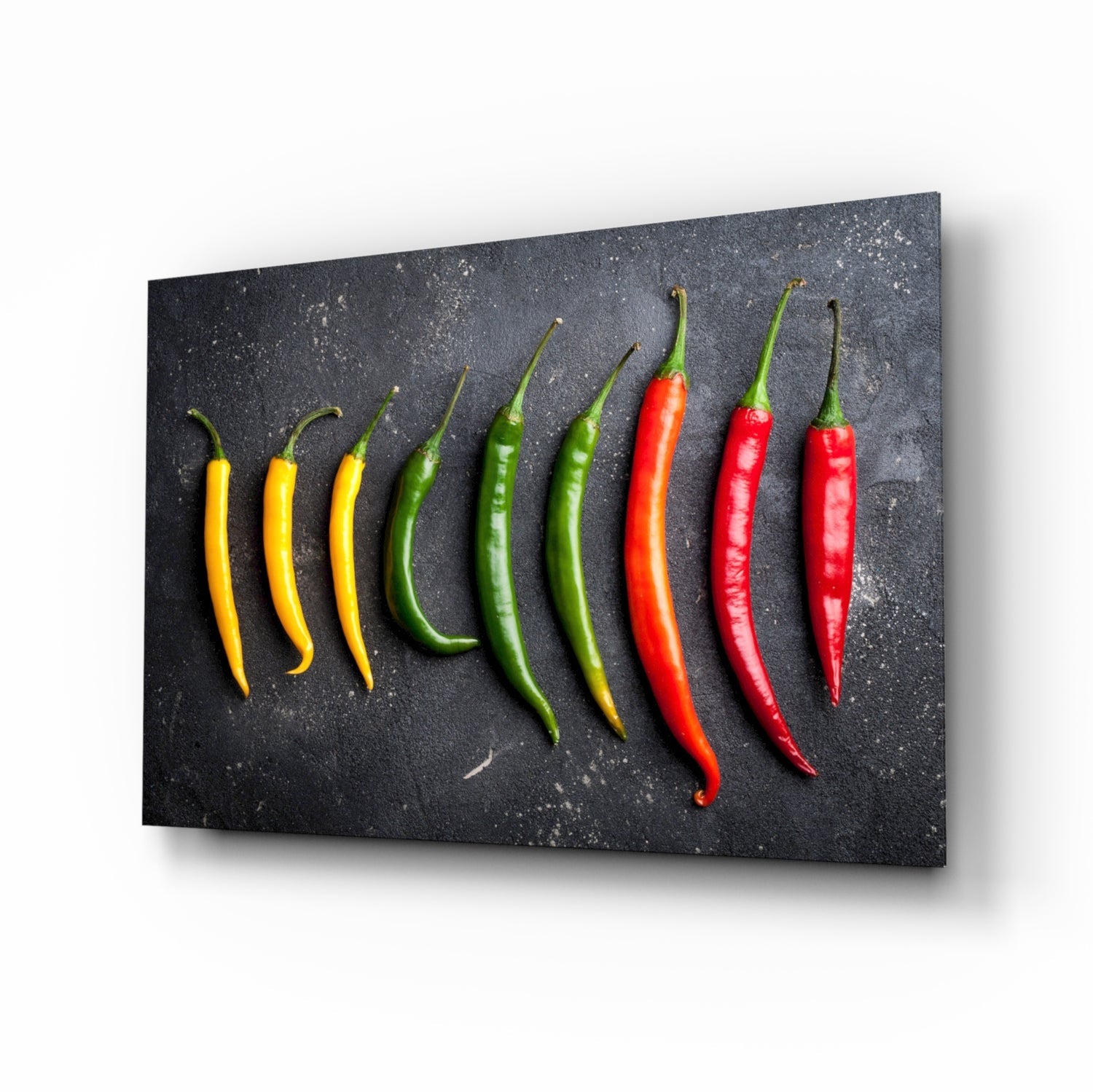 Nine chili peppers in graduating color progression from yellow to green to red arranged in a row on a dark speckled glass panel.