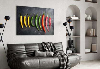 Pepper glass wall art displaying yellow, green, and red chili peppers on dark background, hung above gray leather sofa in modern living room.