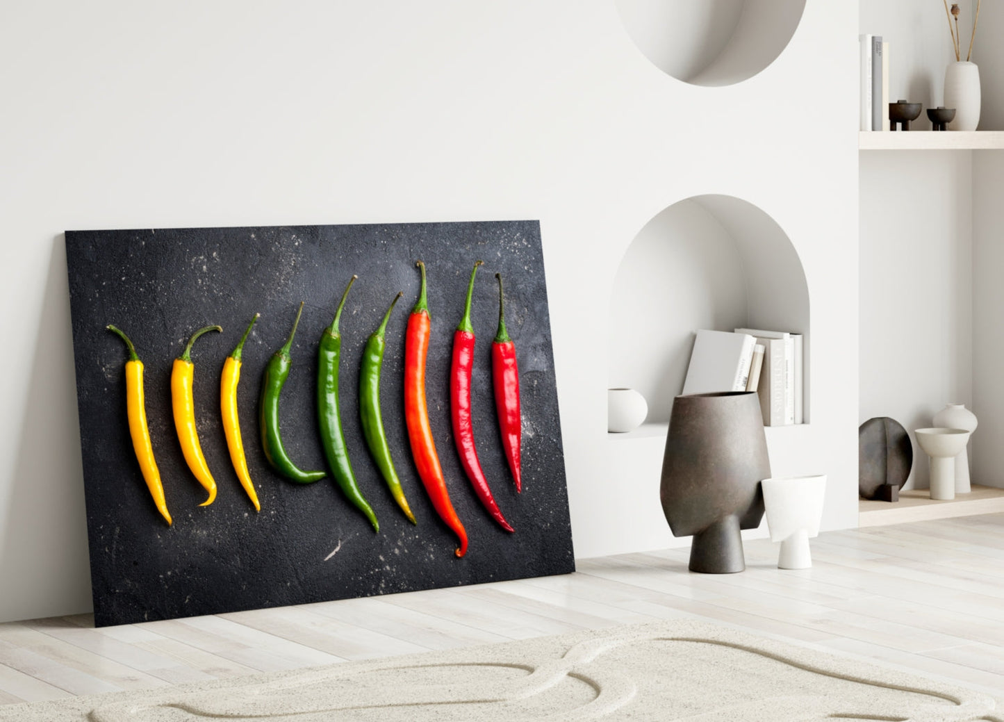 Black glass wall art displaying nine chili peppers in yellow, green, and red gradient, leaning against white wall with decorative objects.