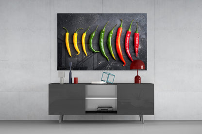 Pepper glass wall art displaying nine chili peppers in yellow, green, and red arranged on black background, mounted above gray media console in modern living room setting.