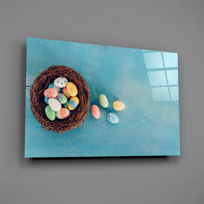 Woven nest filled with colorful painted eggs scattered on turquoise surface with window shadow, displayed at an angle against gray wall.