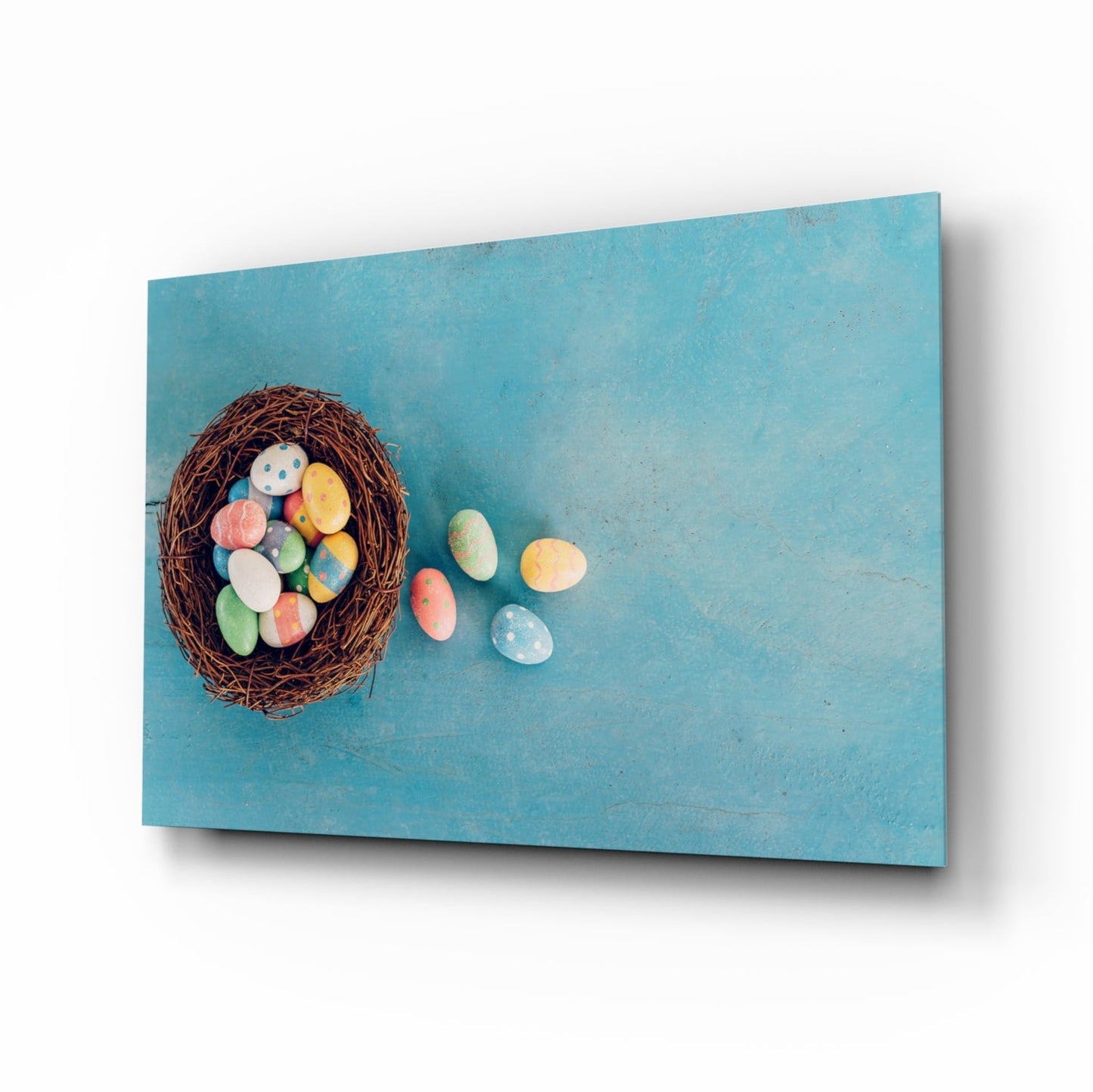 Angled view of turquoise glass wall art displaying a woven nest filled with colorful painted Easter eggs, with several eggs scattered beside it.
