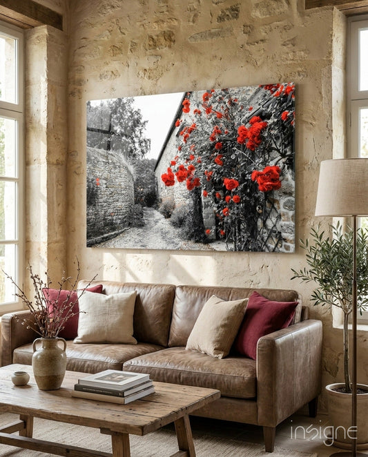 Glass wall art displaying black and white stone alley with vibrant red roses, hung above beige sofa in rustic interior.