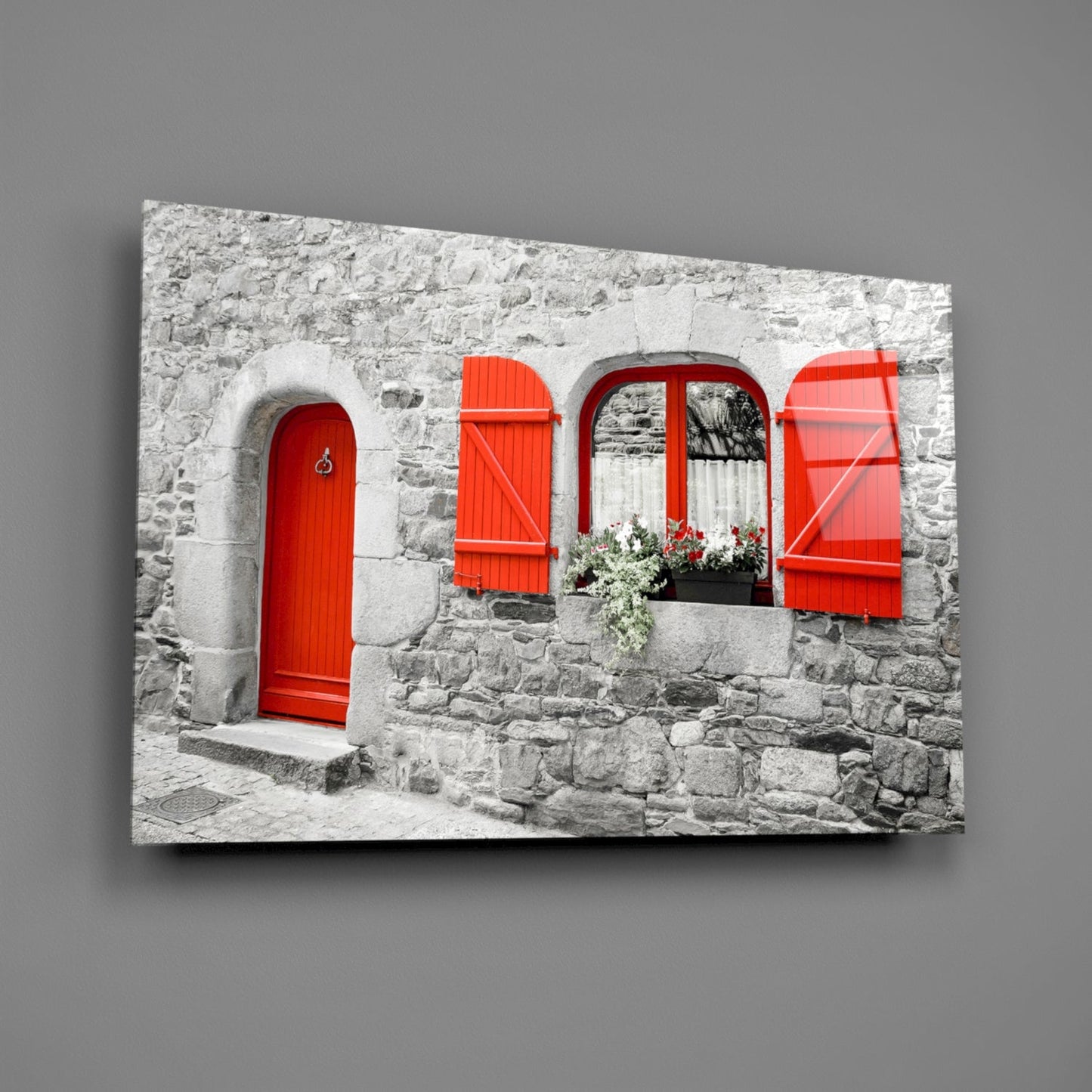 Glass wall art displaying a stone cottage facade with bright red door, shutters, and window box against gray wall.