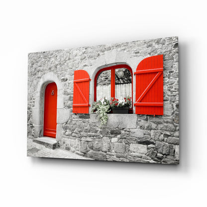 Canvas print displayed at an angle showing a gray stone cottage facade with bright red door, shutters, and flowering window box.