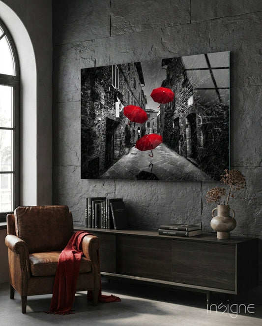 Glass wall art depicting a black and white rainy European street with three red umbrellas, displayed above a dark console table in a modern interior.