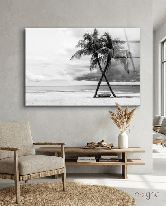 Black and white framed glass art depicting two intertwined palm trees on a beach with ocean and sky backdrop, hung above wooden console table.
