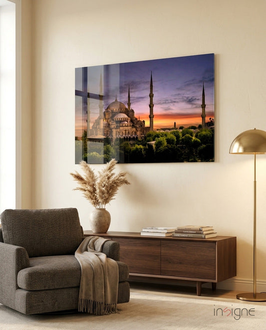 Glass wall art depicting a mosque with purple sunset sky hanging above a wooden console table in a modern living room setting.