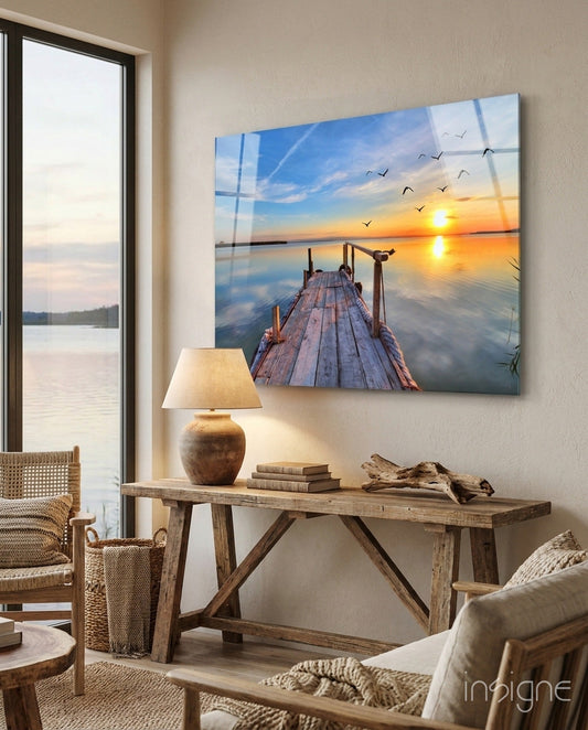 Glass wall art depicting wooden dock at sunset with flying birds, displayed above wooden console table in coastal room.