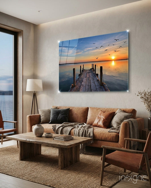 Illuminated glass wall art depicting wooden pier extending toward sunset over water with flying birds, displayed above brown leather sofa in modern living room.