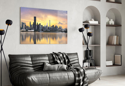 Horizontal glass wall art depicting a golden-hour Manhattan skyline with water reflection, displayed above a black leather sofa in a modern living room.