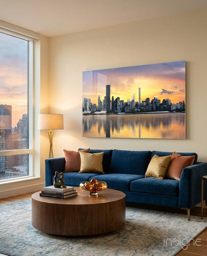 Downtown Glass Wall Art displaying a sunset cityscape with golden sky reflected in water, mounted above a blue sofa in a modern living room.