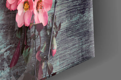 Three-dimensional angled view of glass wall art displaying pink flowers with yellow stamens against weathered teal wooden texture background.