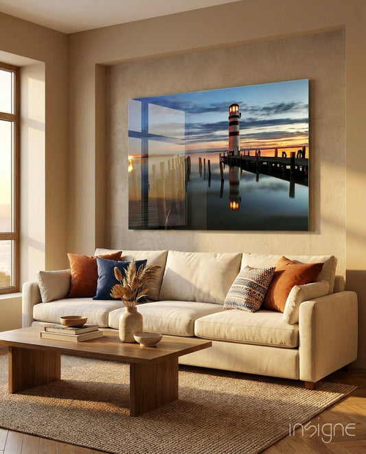 Glass wall art depicting a striped lighthouse at sunset reflected in calm water, displayed above a cream sofa in a warm, neutral living room.