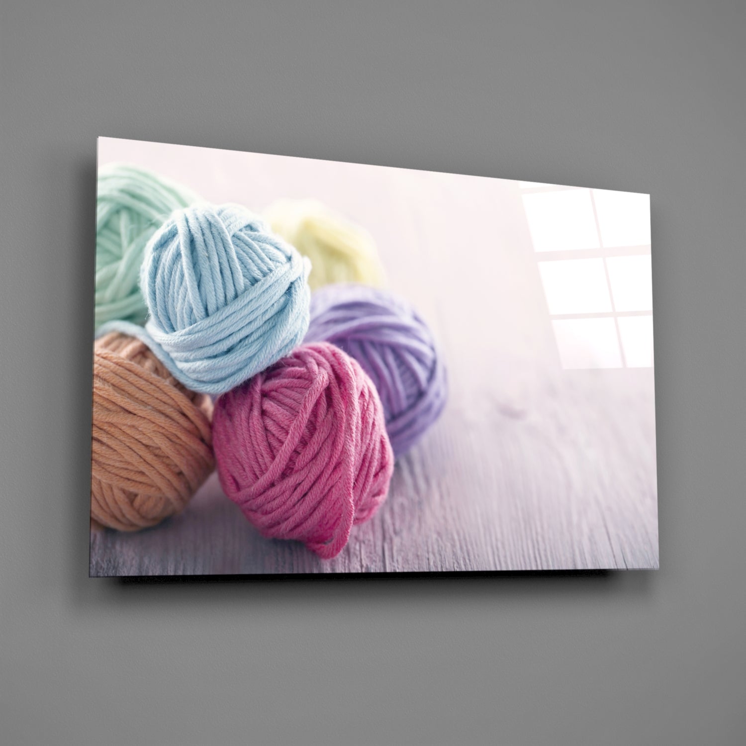 Colorful yarn balls in blue, pink, purple, tan, and green arranged on white wooden surface with window background, displayed as glass wall art.