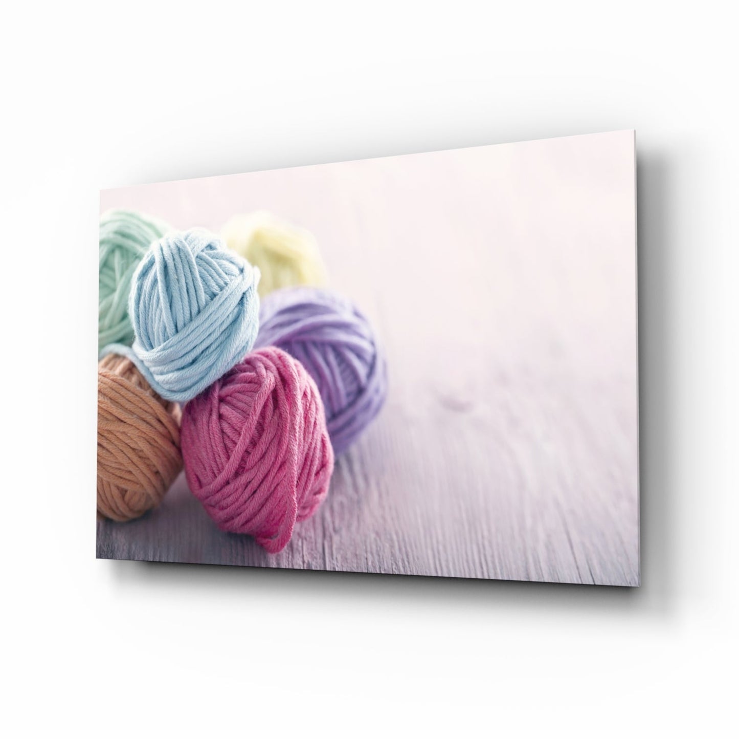 Colorful yarn balls in blue, pink, purple, and brown arranged on white wooden surface, displayed as glass wall art at an angle.