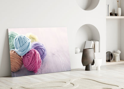 Colorful yarn balls in blue, pink, purple, and tan displayed on glass wall art leaning against white interior wall.