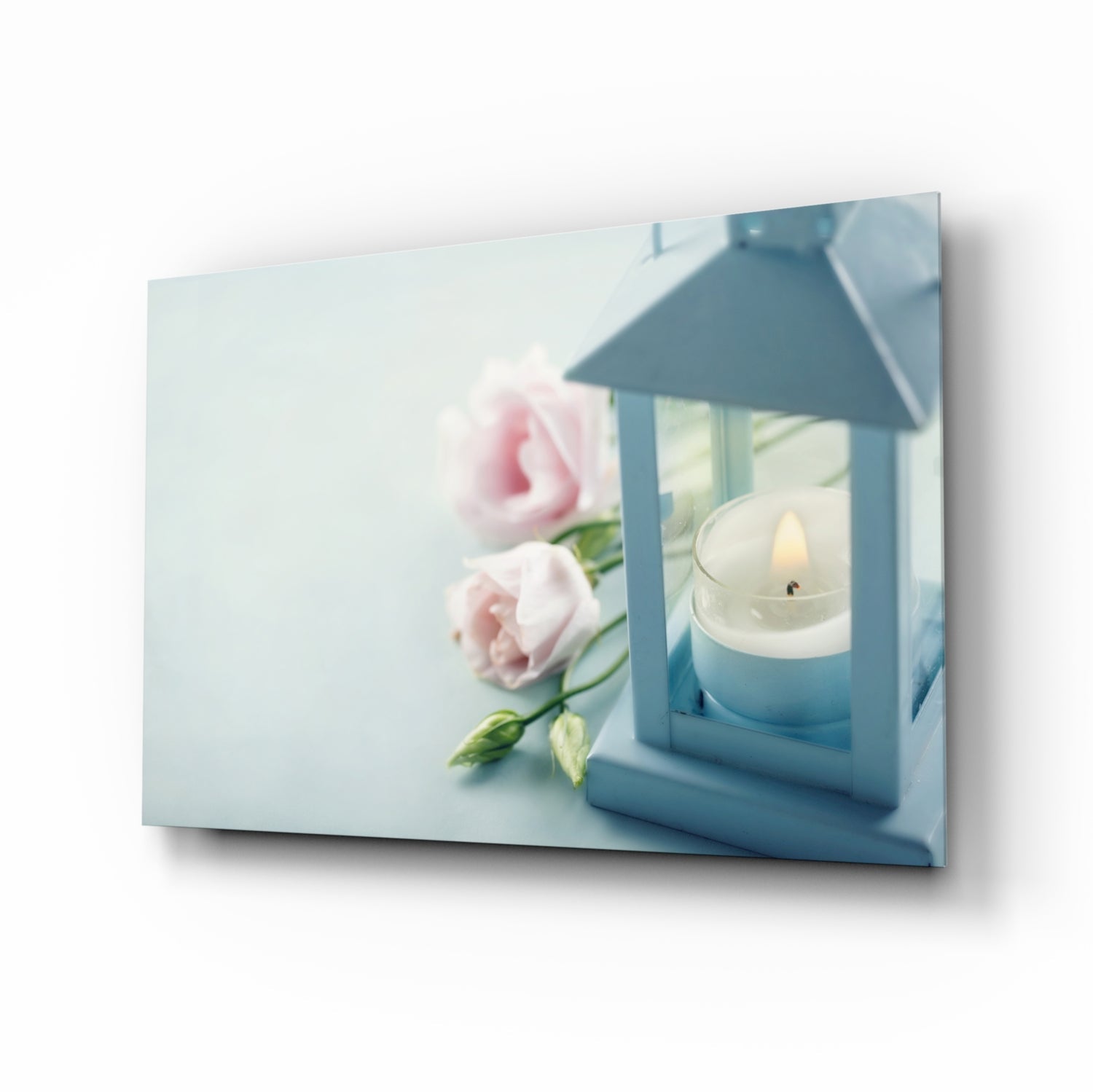 Glass wall art displaying pink roses beside a blue lantern with lit white candle, angled perspective view.
