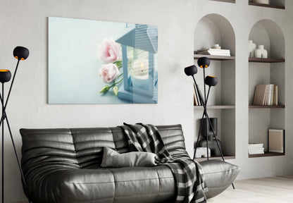 Horizontal glass wall art depicting pink roses and blue lantern with candle, displayed above black leather sofa in modern living room.