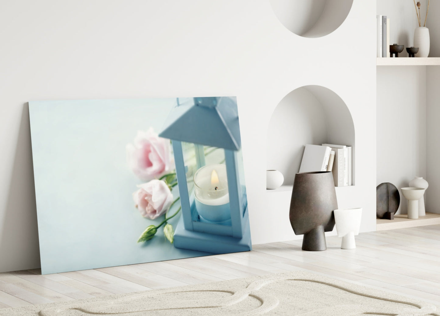 Light blue glass canvas leaning against white wall displays pink roses and blue lantern with candle beside minimalist home décor objects.