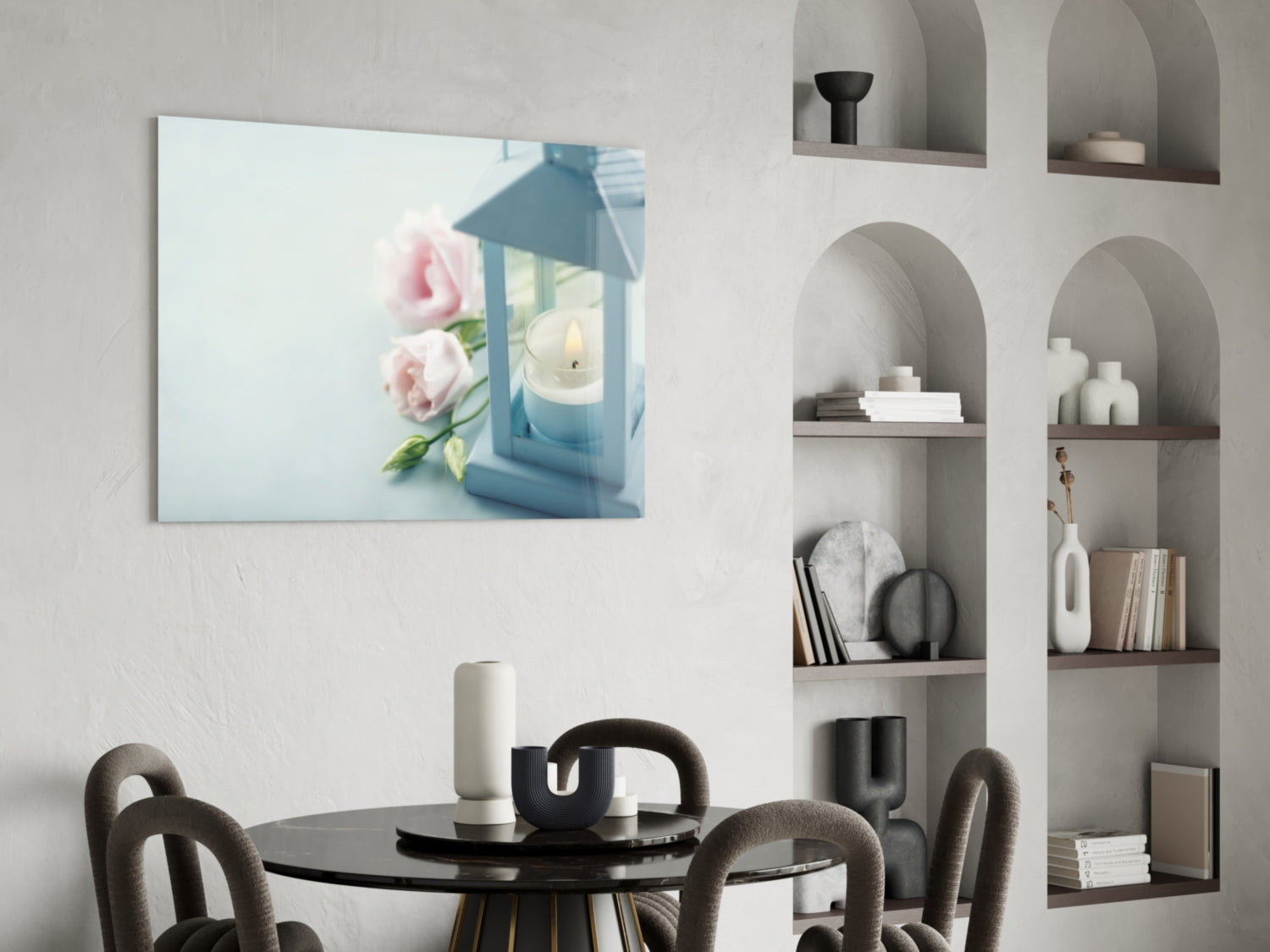 Glass wall art featuring pink roses and blue lantern with candle displayed above round black dining table in minimalist interior.
