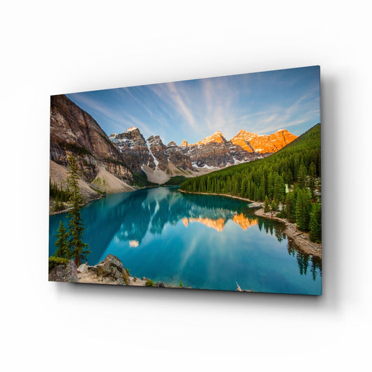 Glass wall art panel displaying a mountain lake landscape with turquoise water, snow-capped peaks, and evergreen forests at sunrise.
