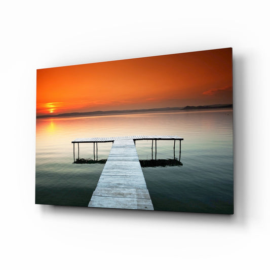 Glass wall art panel displaying a wooden dock extending into calm water at sunset, with orange sky and silhouetted boathouses.
