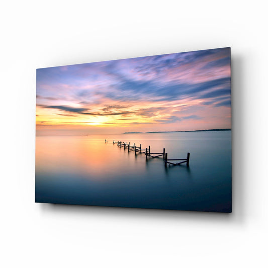 Glass wall art panel displaying a serene sunset over calm water with wooden pier posts receding into the horizon, mounted at an angle showing shadow and depth.