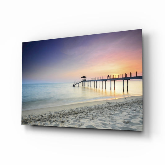 Glass wall art panel displaying a serene beach sunrise with wooden pier, soft sand, and pastel sky gradient, mounted at an angle.
