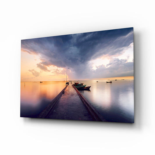 Long wooden pier extending into calm water at sunset with moored fishing boats and dramatic cloudy sky above, displayed as glass wall art.